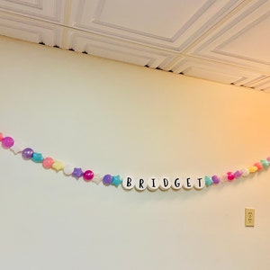 May include: A white wall with a colourful garland spelling out the name "BRIDGET". The garland is made of white plastic letters, colourful plastic balls, and stars.