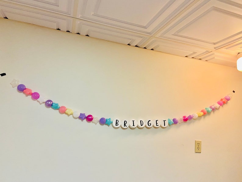 May include: A white wall with a colourful garland spelling out the name "BRIDGET". The garland is made of white plastic letters, colourful plastic balls, and stars.