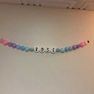 May include: A pastel-coloured beaded garland with the name "ROSE" spelled out in white letters. The garland is strung with pink, purple, and blue beads.