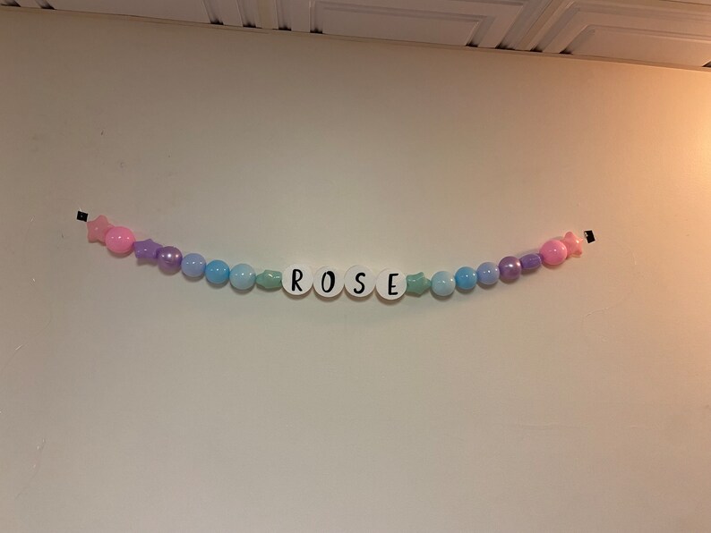 May include: A pastel-coloured beaded garland with the name "ROSE" spelled out in white letters. The garland is strung with pink, purple, and blue beads.