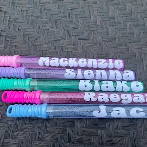 May include: Five bubble wands with names in white script: Mackenzie, Sienna, Blake, Raegan, and Jack. The wands have colorful handles in pink, purple, green, pink, and blue. The tubes are clear and filled with bubble solution.