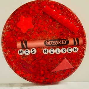May include: Red glitter resin coaster with a red crayon, a star, a paperclip, and a triangle. The coaster has the text "Crayola" and "Mrs. Nelsen" in white letters.