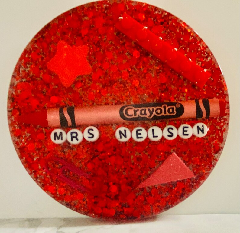 May include: Red glitter resin coaster with a red crayon, a star, a paperclip, and a triangle. The coaster has the text "Crayola" and "Mrs. Nelsen" in white letters.
