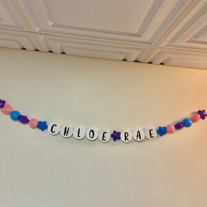 May include: A white banner with the name "CHLOE RAE" spelled out in black letters. The banner is decorated with purple stars and pink and blue balloons.