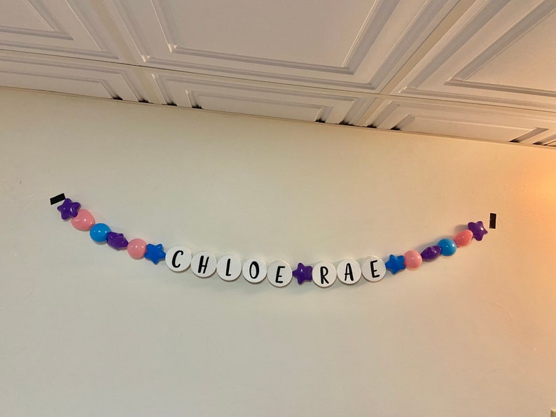 May include: A white banner with the name "CHLOE RAE" spelled out in black letters. The banner is decorated with purple stars and pink and blue balloons.