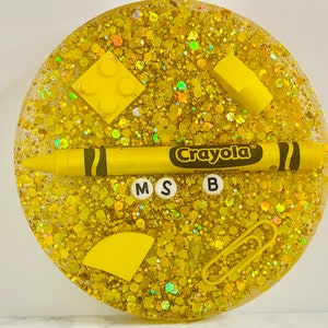 May include: A yellow glitter resin coaster with a Crayola crayon, a yellow Lego brick, a yellow semi-circle, a yellow rectangle, a paperclip, and the letters M, S, and B.
