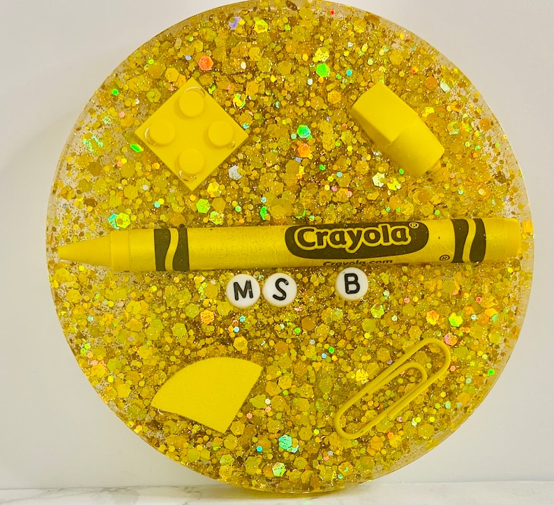 May include: A yellow glitter resin coaster with a Crayola crayon, a yellow Lego brick, a yellow semi-circle, a yellow rectangle, a paperclip, and the letters M, S, and B.