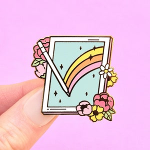 May include: Gold enamel pin featuring a stylised drawing of a tablet with a rainbow and a stylus. The tablet is surrounded by pink, yellow and white flowers.