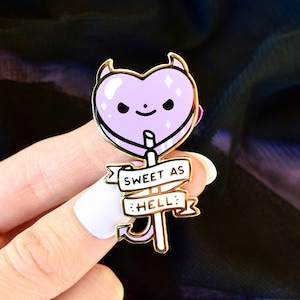 Puede incluir: Un pin de esmalte en forma de corazón lavanda con cuernos de diablo y una cara linda. El pin tiene un borde dorado y una pancarta que dice "SWEET AS HELL". El pin está sostenido por una mano.