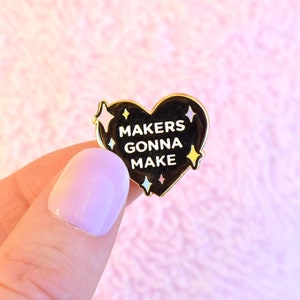 May include: Gold heart-shaped enamel pin with black background and white text that reads "Makers Gonna Make". The pin is decorated with small gold, pink, and blue stars.