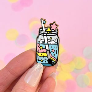 Puede incluir: Un pin de esmalte dorado con un tarro de cristal lleno de remolinos azules, rosas y amarillos, estrellas y un pincel blanco.