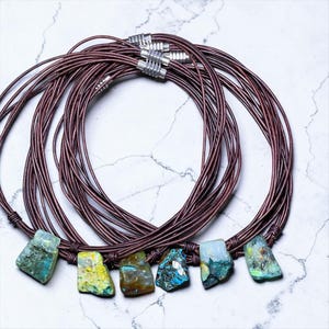 May include: A set of necklaces featuring multiple dark brown leather cords and silver-toned cylindrical clasps. Each necklace is adorned with a single raw, irregularly shaped gemstone pendant in varying shades of blue, green, and yellow.