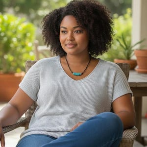 May include: A light gray short-sleeved V-neck sweater with a textured knit pattern, paired with blue denim jeans and a necklace featuring a black cord and a series of small, cylindrical turquoise-colored beads centered at the neckline.