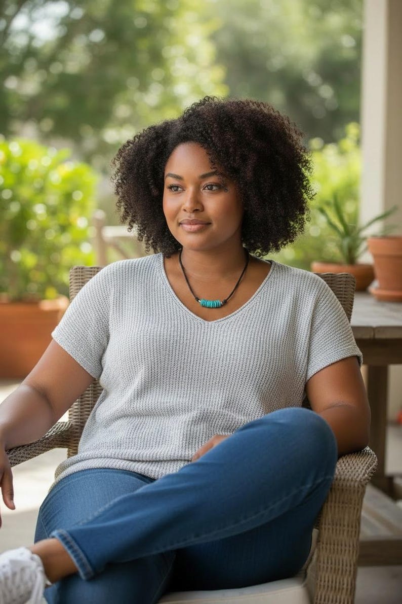 May include: A light gray short-sleeved V-neck sweater with a textured knit pattern, paired with blue denim jeans and a necklace featuring a black cord and a series of small, cylindrical turquoise-colored beads centered at the neckline.