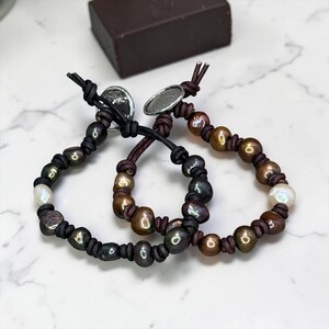 May include: Two bracelets crafted from dark brown leather cord featuring a series of iridescent, multi-colored freshwater pearls in shades of bronze, charcoal, and white. Each bracelet is secured with a round, metallic silver-toned button clasp.