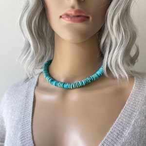 May include: A turquoise blue beaded necklace made of round, flat beads. The necklace is worn by a model with silver-grey hair.
