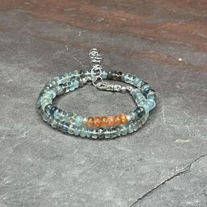 May include: A delicate necklace made with light blue and gray gemstone beads, accented with a row of orange beads in the center. The necklace has a silver clasp.