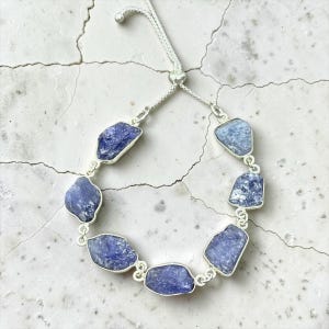 May include: A silver adjustable bracelet featuring seven raw, uncut blue tanzanite stones set in silver bezels. The bracelet includes a sliding bead mechanism with thin silver chains and small silver spheres at the ends to secure the fit.