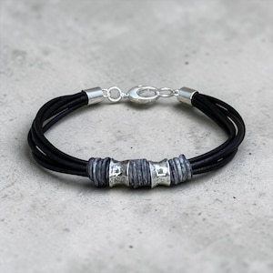 May include: A multi-strand black leather cord bracelet featuring a silver-toned lobster claw clasp and a central arrangement of two hammered silver-toned metal beads alternating with three dark grey textured cylindrical spacer beads.