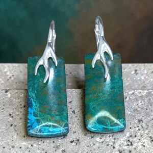 May include: A pair of rectangular earrings featuring polished teal and brown patterned stone slabs. Each stone is topped with a silver-toned metal accent shaped like a small, branching antler or twig, connected to a curved ear wire hook.