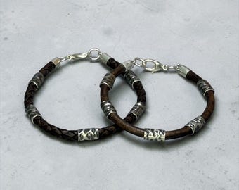 Op de afbeelding: Twee armbanden met donkerbruine leren koorden, één gevlochten en één glad, geaccentueerd met zilverkleurige gehamerde metalen cilindrische kralen en afgewerkt met zilverkleurige karabijnsluitingen.