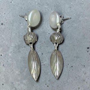 May include: A pair of drop earrings featuring a smooth, round, off-white pearl stud at the top, connected to a textured, silver-toned metal bead with a leaf pattern, and finished with a long, pointed, silver-toned metal leaf-shaped pendant.