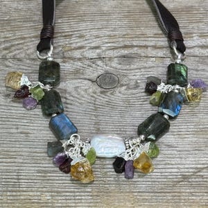 May include: A handmade necklace with a selection of colourful gemstones, such as labradorite, citrine, and amethyst. The necklace has a dark brown leather cord and silver-toned metal details. The gemstones are different sizes and shapes.