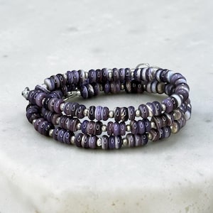May include: A multi-strand wrap bracelet featuring small, disc-shaped purple and white marbled beads alternating with tiny, round, metallic silver-toned spacer beads. The bracelet is coiled in a circular shape, showcasing a consistent pattern.