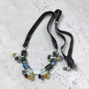 May include: A necklace featuring a dark brown leather cord and a collection of multi-colored gemstone beads, including labradorite, amethyst, and citrine, accented with silver-toned metal findings and a central white iridescent oval bead.