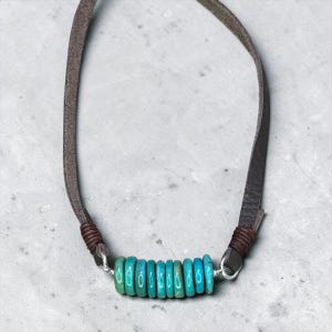 May include: A necklace featuring a row of eight small, round, turquoise-colored beads strung on a silver wire. The beads are flanked by dark brown leather cord sections, which are secured with dark brown thread wraps at each end of the wire.