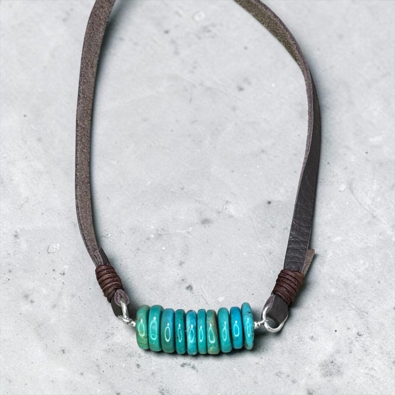 May include: A necklace featuring a row of eight small, round, turquoise-colored beads strung on a silver wire. The beads are flanked by dark brown leather cord sections, which are secured with dark brown thread wraps at each end of the wire.