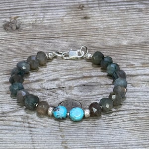 May include: A beaded bracelet featuring faceted gray-green beads and two turquoise beads. The bracelet has a silver clasp and is displayed on a wooden surface. The bracelet is a piece of jewelry.