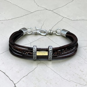 Puede incluir: Una pulsera de múltiples hilos con cordones de cuero marrón oscuro y un hilo de cuero trenzado. El diseño incluye dos espaciadores de metal plateado texturizado que flanquean una barra rectangular dorada lisa, con un cierre de gancho plateado.