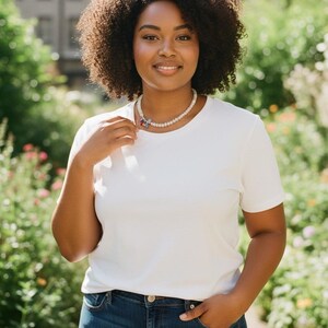 K&ouml;nnte beinhalten: Ein wei&szlig;es kurz&auml;rmeliges T-Shirt mit Rundhalsausschnitt, kombiniert mit einer dunkelblauen Jeans und einer Perlenkette mit einem kleinen, mehrfarbigen Blumenanh&auml;nger. Die Kette liegt am Halsansatz und das T-Shirt hat ein schlichtes, einfarbiges Design.