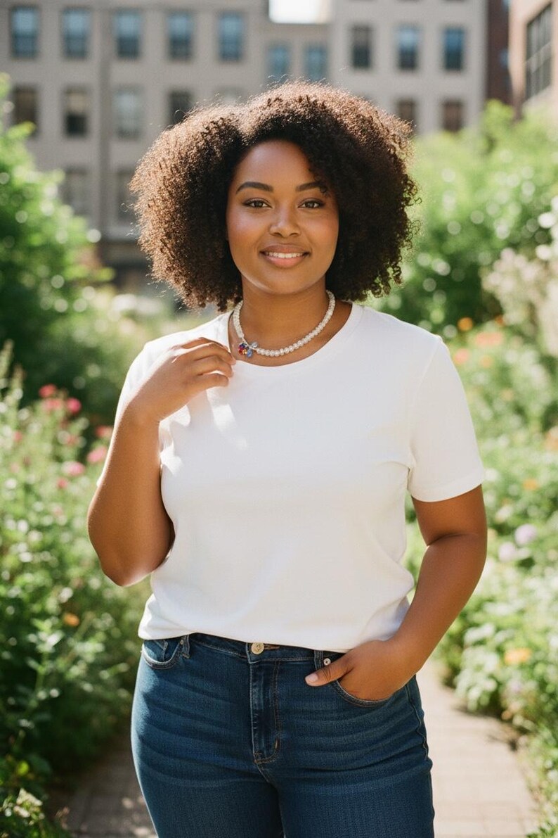 K&ouml;nnte beinhalten: Ein wei&szlig;es kurz&auml;rmeliges T-Shirt mit Rundhalsausschnitt, kombiniert mit einer dunkelblauen Jeans und einer Perlenkette mit einem kleinen, mehrfarbigen Blumenanh&auml;nger. Die Kette liegt am Halsansatz und das T-Shirt hat ein schlichtes, einfarbiges Design.