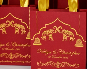 May include: Red gift bags with gold handles and gold elephant designs. The bags have the text "Nithya & Christopher" and "30 December 2022" printed on them. The bags also have the text "Thank you for celebrating our special day with us!"