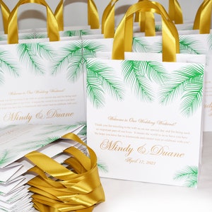 May include: White favor bags with gold ribbon handles and a green palm tree design. The bags have the text "Welcome to Our Wedding Weekend!" and "Mindy & Duane" with the date "April 17, 2021".