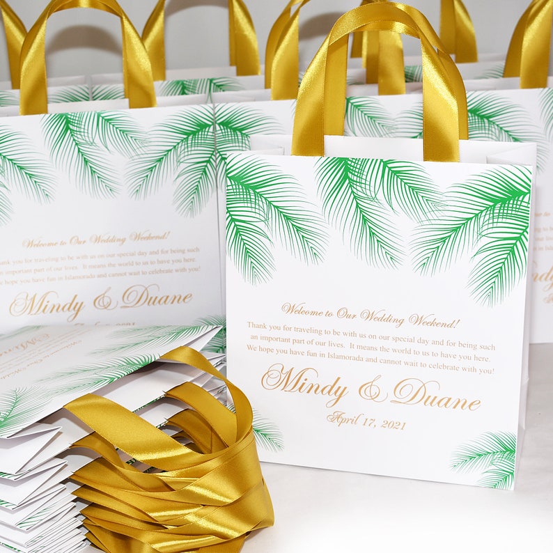 May include: White favor bags with gold ribbon handles and a green palm tree design. The bags have the text "Welcome to Our Wedding Weekend!" and "Mindy & Duane" with the date "April 17, 2021".