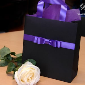 May include: A black gift bag with a purple ribbon and bow. The bag is filled with purple tissue paper. A white rose is in the foreground.