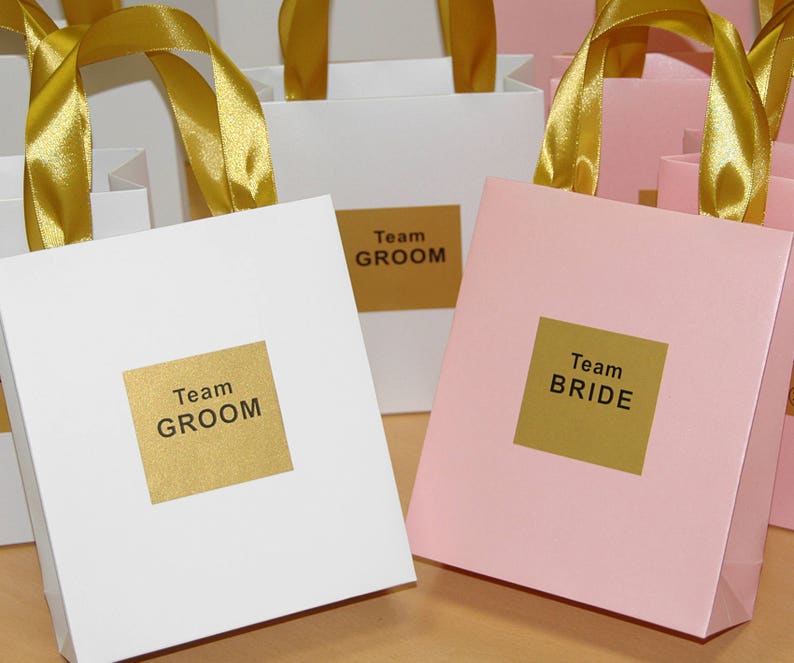 May include: White and pink gift bags with gold ribbon handles. The bags have gold labels with the text "Team Groom" and "Team Bride".