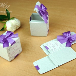 May include: Three white favor boxes with purple ribbon bows and a personalized label that reads "Violetta & Erik!"  The boxes are made of a textured paper and are perfect for holding small gifts or treats.