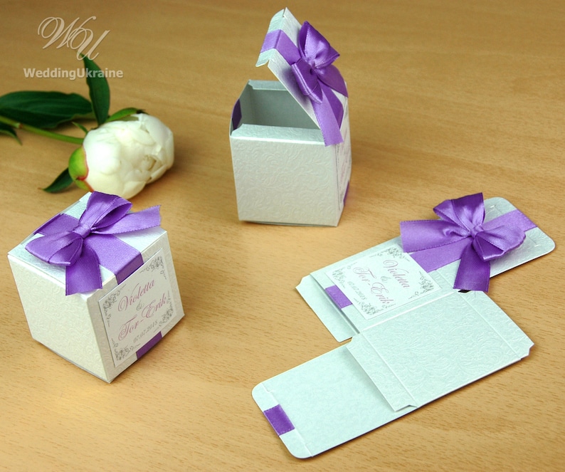 May include: Three white favor boxes with purple ribbon bows and a personalized label that reads "Violetta & Erik!"  The boxes are made of a textured paper and are perfect for holding small gifts or treats.