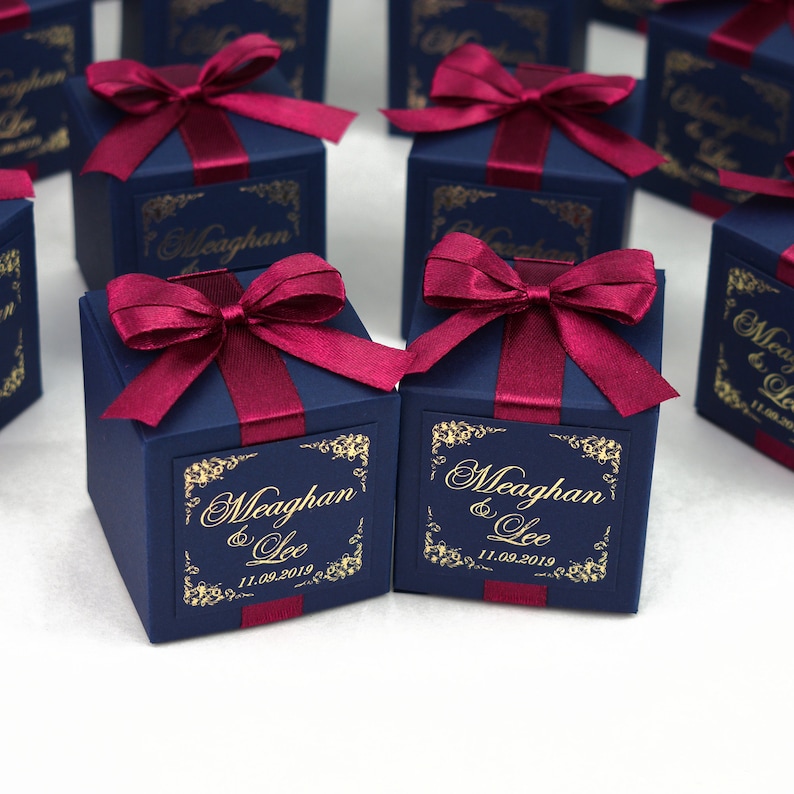 May include: Dark blue gift boxes tied with burgundy ribbon bows. Each box features a gold label with the names "Meaghan & Lee" and the date "11.09.2019". The boxes are square and set on a white surface.