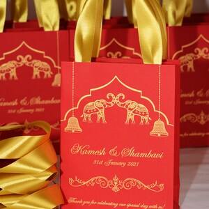 Op de afbeelding: Rode cadeauzakjes met gouden lintjes en gouden olifantmotieven. De zakjes hebben de tekst "Kamesh & Shambavi 31st January 2021" en "Bedankt dat je onze speciale dag met ons hebt gevierd!"