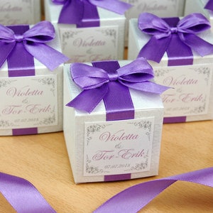 May include: White favor boxes with purple ribbon bows and personalized labels. The labels read "Violetta & Tor-Erik" and "07.07.2018".