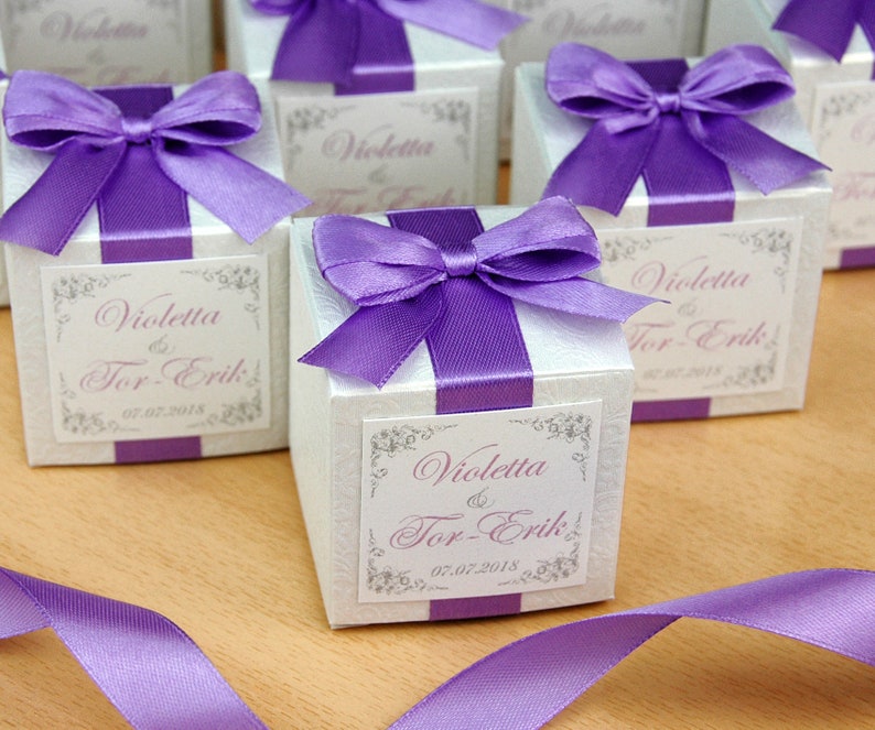 May include: White favor boxes with purple ribbon bows and personalized labels. The labels read "Violetta & Tor-Erik" and "07.07.2018".