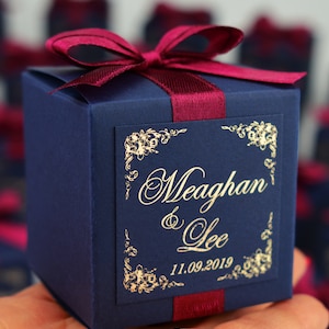 May include: A navy blue gift box tied with a burgundy ribbon and bow. The box features a gold-framed label with the names "Meaghan & Lee" and the date "11.09.2019". The box is held in a hand, with other similar boxes out of focus in the background.