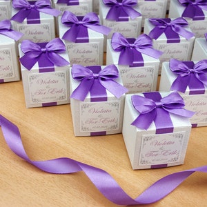 May include: A collection of small white gift boxes with purple satin ribbon bows. Each box has a label with the text "Violetta & Tor-Erik" and the date "07.07.2018".