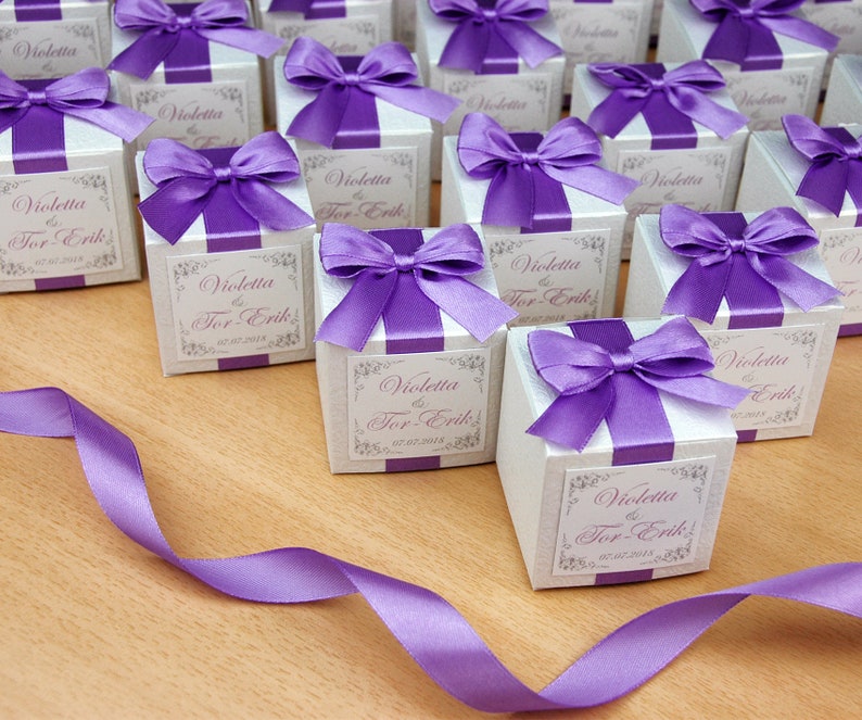 May include: A collection of small white gift boxes with purple satin ribbon bows. Each box has a label with the text "Violetta & Tor-Erik" and the date "07.07.2018".