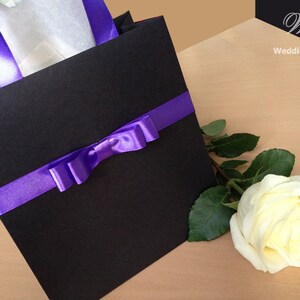 May include: A black gift bag with a purple ribbon and bow. A single white rose with green leaves is next to the bag.
